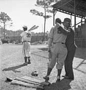 Two white men in baseball uniform with back to camera watch a black 
